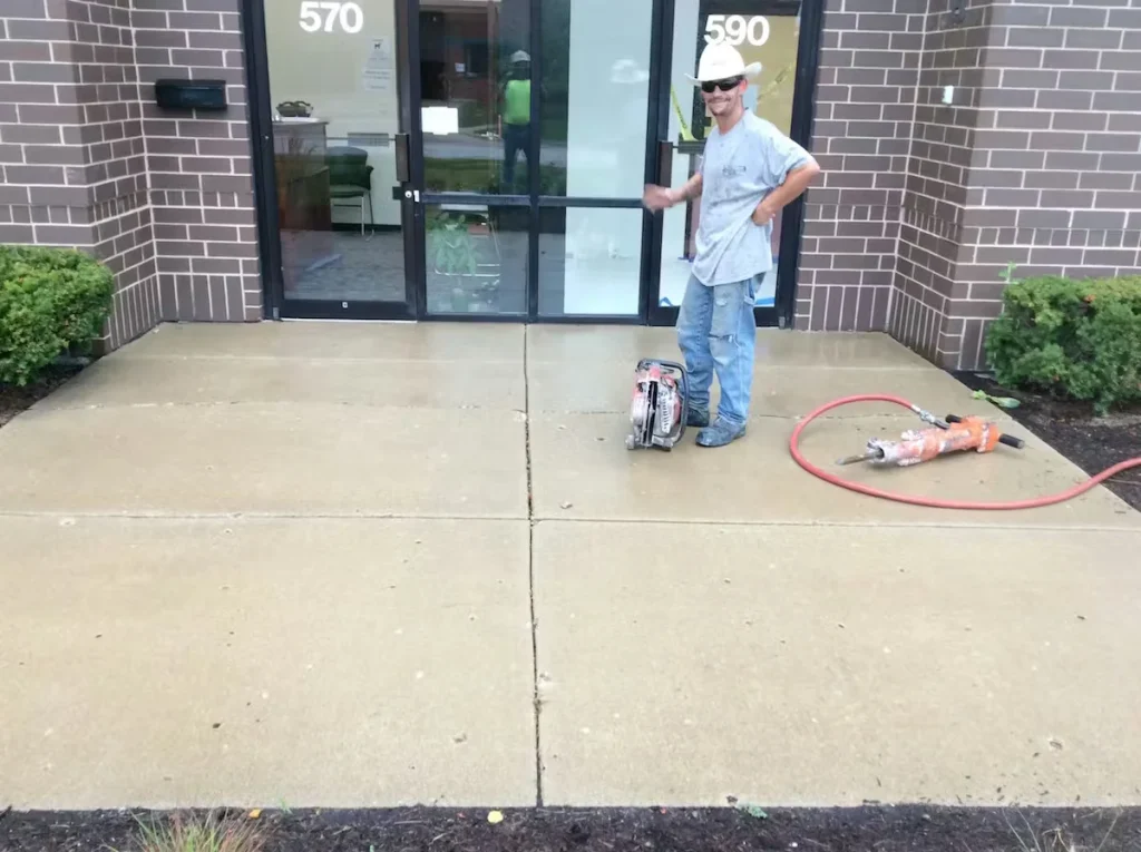 Worker on site during a stamped concrete installation, standing on freshly prepared concrete in front of a commercial office entrance with cutting and finishing tools staged nearby.