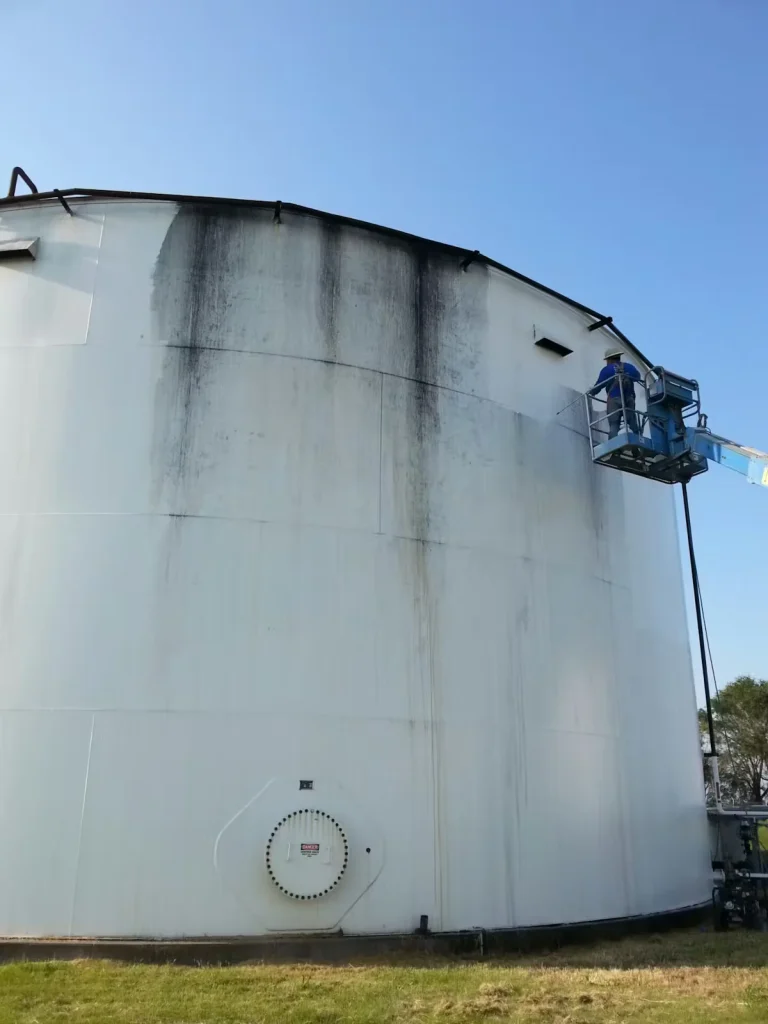 Industrial storage tank exterior with visible staining as a worker uses a boom lift to perform power washing, sandblasting, and surface preparation before repainting
