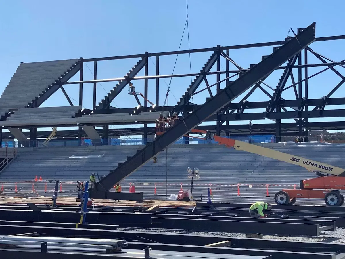Steel stadium framework under construction with workers performing touch-up painting using boom lifts and scaffolding at an outdoor construction site
