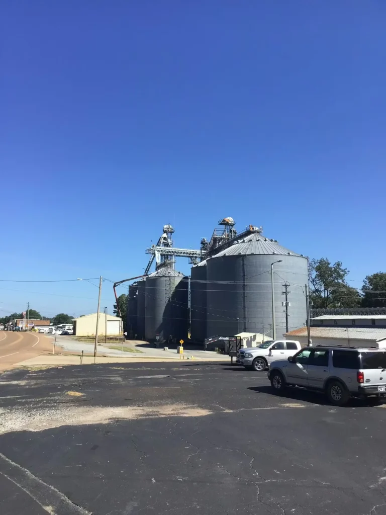 Industrial grain silos and processing structures at a food production facility with parked vehicles and clear blue sky