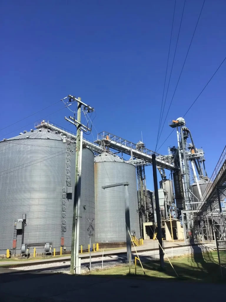 Closer view of large metal silos, conveyors, and support towers at an industrial processing facility with utility lines in the foreground