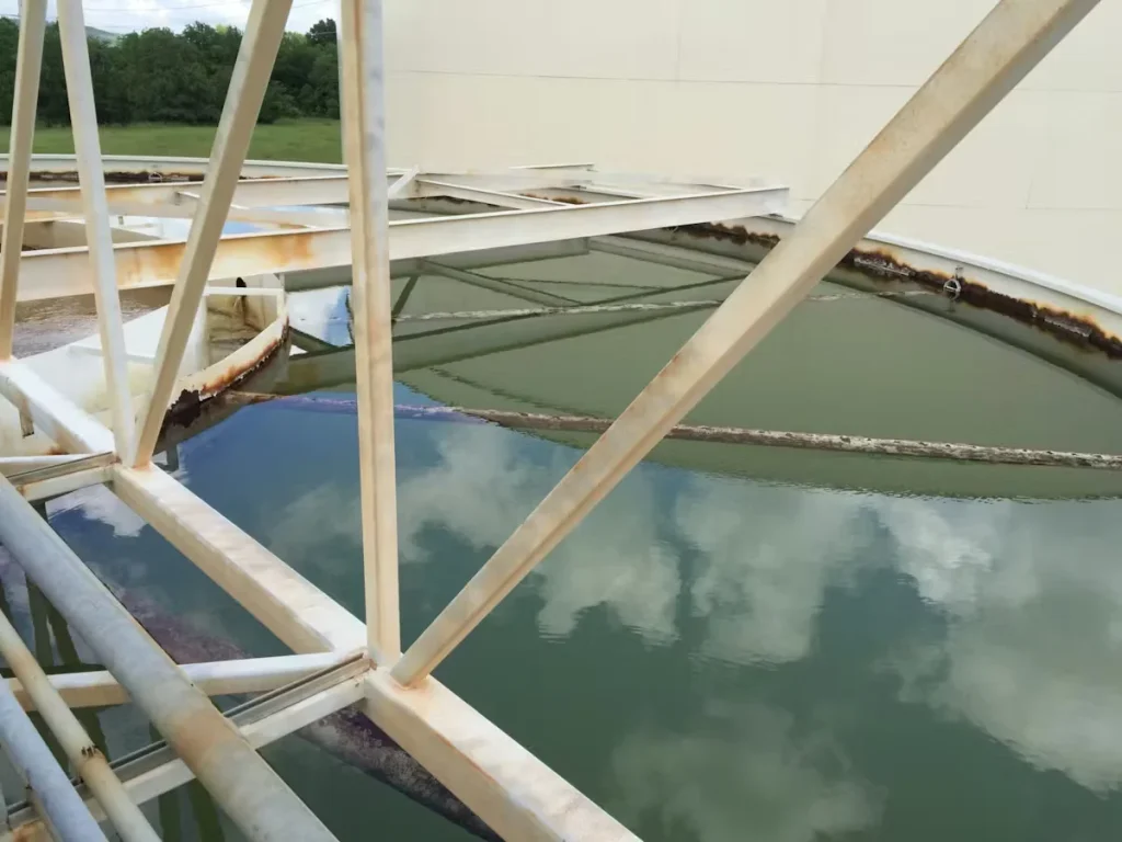 Industrial steel structure above water basin showing corrosion and surface preparation prior to vapor-assisted sandblasting and repainting