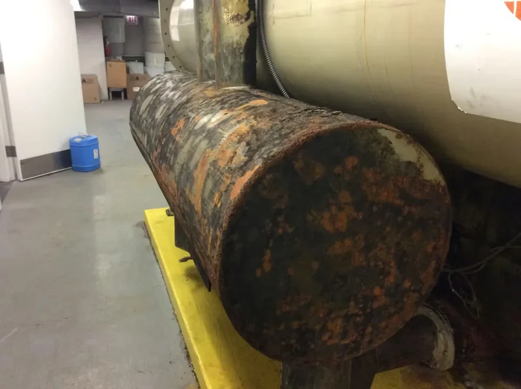 Corroded industrial steel tank and piping with heavy rust buildup prior to vapor-assisted sandblasting and urethane coating application