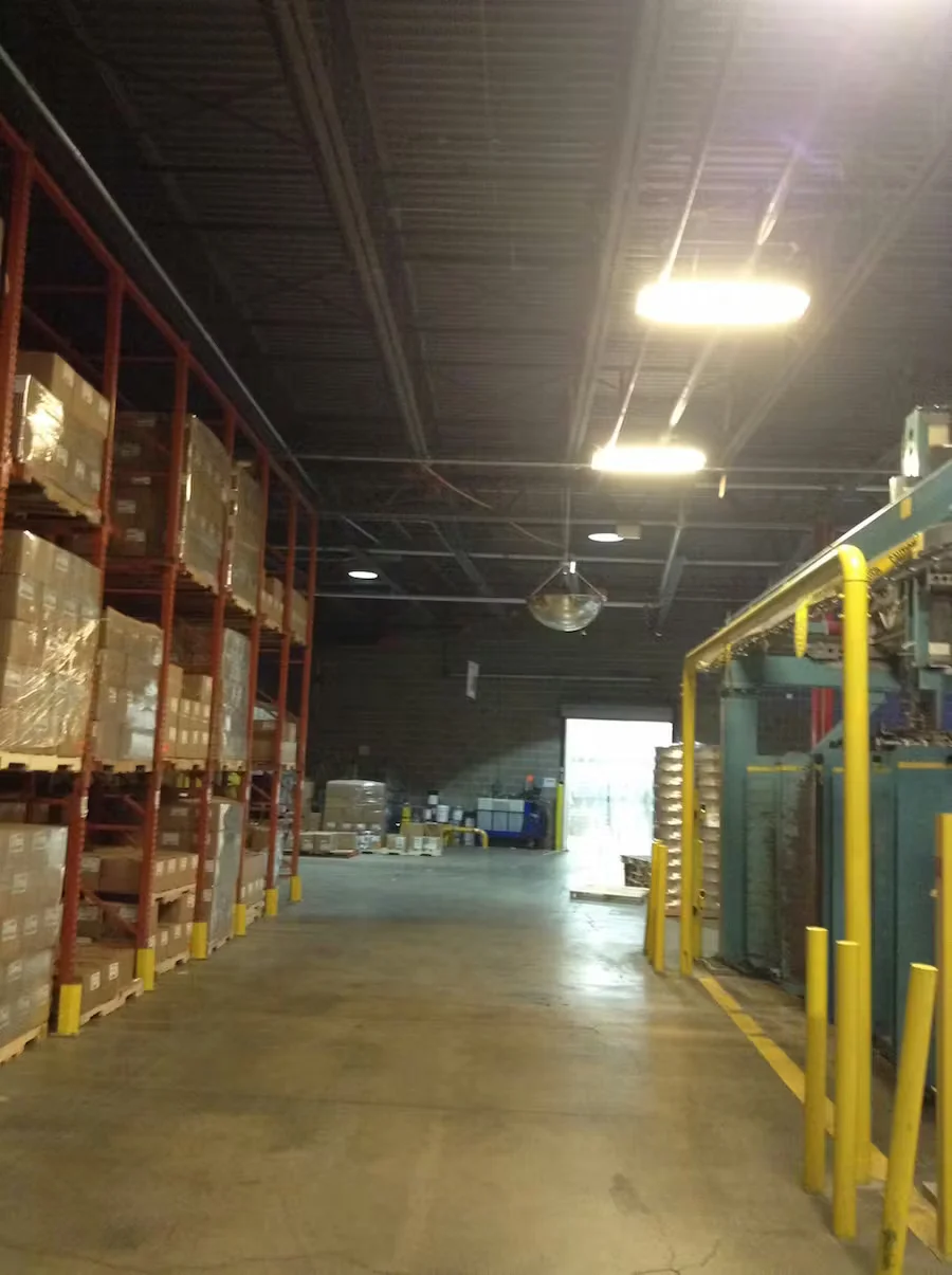 Warehouse interior aisle with tall pallet racking, industrial machinery, overhead lighting, and exposed ceiling before painting