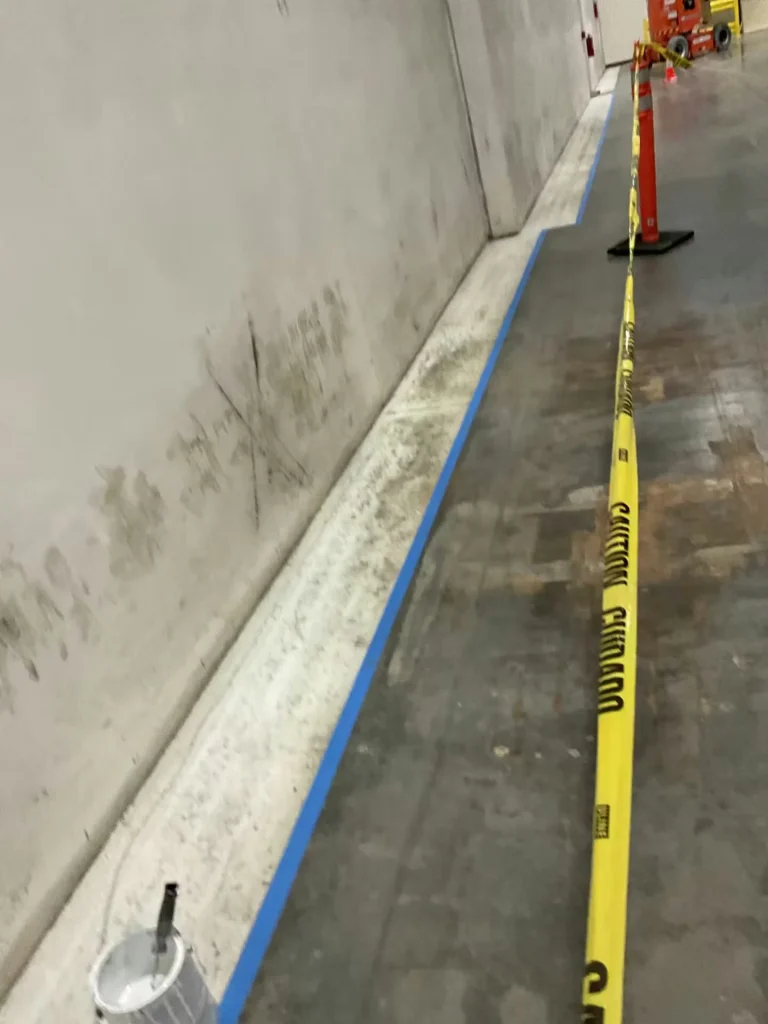 Warehouse floor edge masked with blue tape and caution tape during epoxy floor preparation along wall base