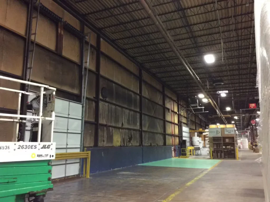 Warehouse interior with high ceilings, industrial lighting, wall insulation panels, and a painted work area on the floor