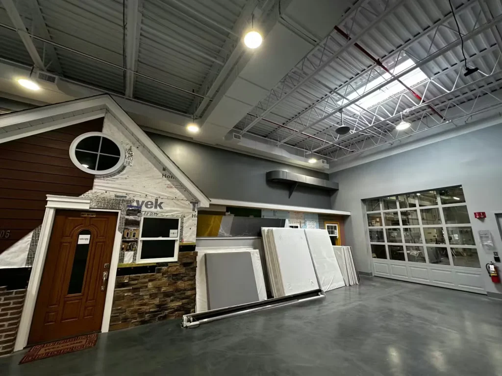 Warehouse showroom displaying building materials, model home exterior, and high industrial ceiling